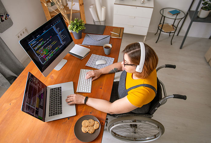 A high-view of a woman in a wheelchair with multiple monitors with graphs and charts on them and a plate of cookies at her desk